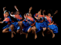 Five women in blue and orange outfits mid-air, performing a joyful dance against a black background