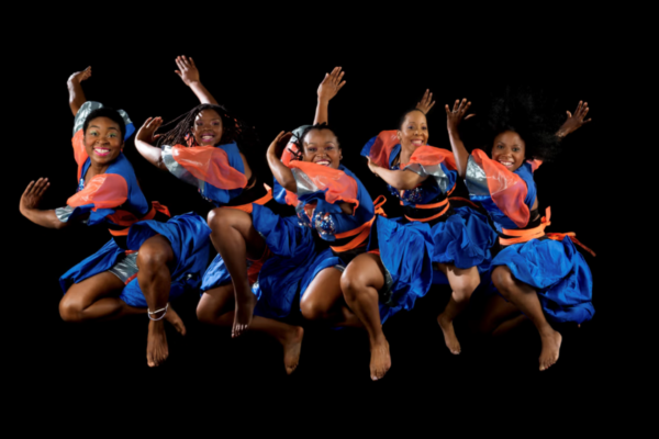 Five women in blue and orange outfits mid-air, performing a joyful dance against a black background