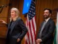 Two officials standing at a podium, smiling, with U.S. flags behind them during a formal event